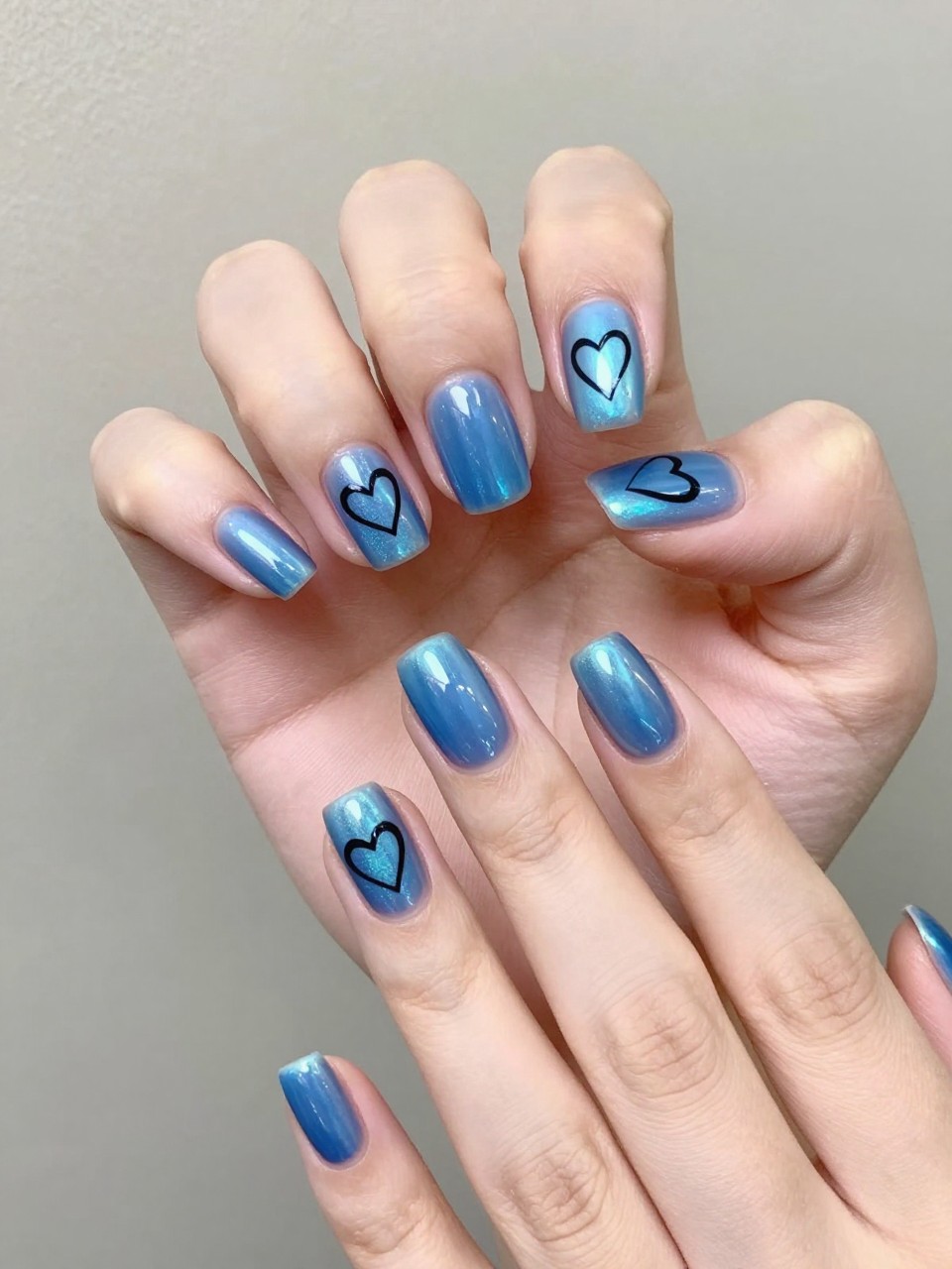 Hands moving under a light to show holographic shift. The nails are a shifting blue holographic. A crisp black heart outline is painted on each nail. The background is neutral to keep focus on the dynamic colors.