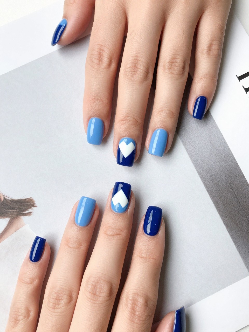 Top-down view of hands with square nails on a modern design magazine. Each nail is split diagonally into powder blue and navy. A geometric white heart is painted at the seam. The look is crisp and fashion-forward.