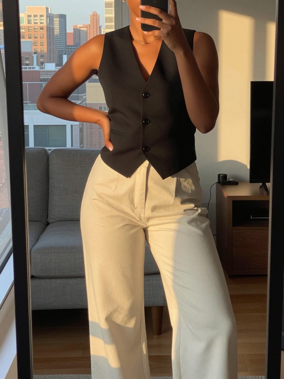 iPhone mirror selfie of a black woman wearing a black vest with cream wide-leg pants, posed with one hand on her hip, cropped at NECK so face doesn't show, in a city apartment, evening golden hour light, iPhone photo quality.
