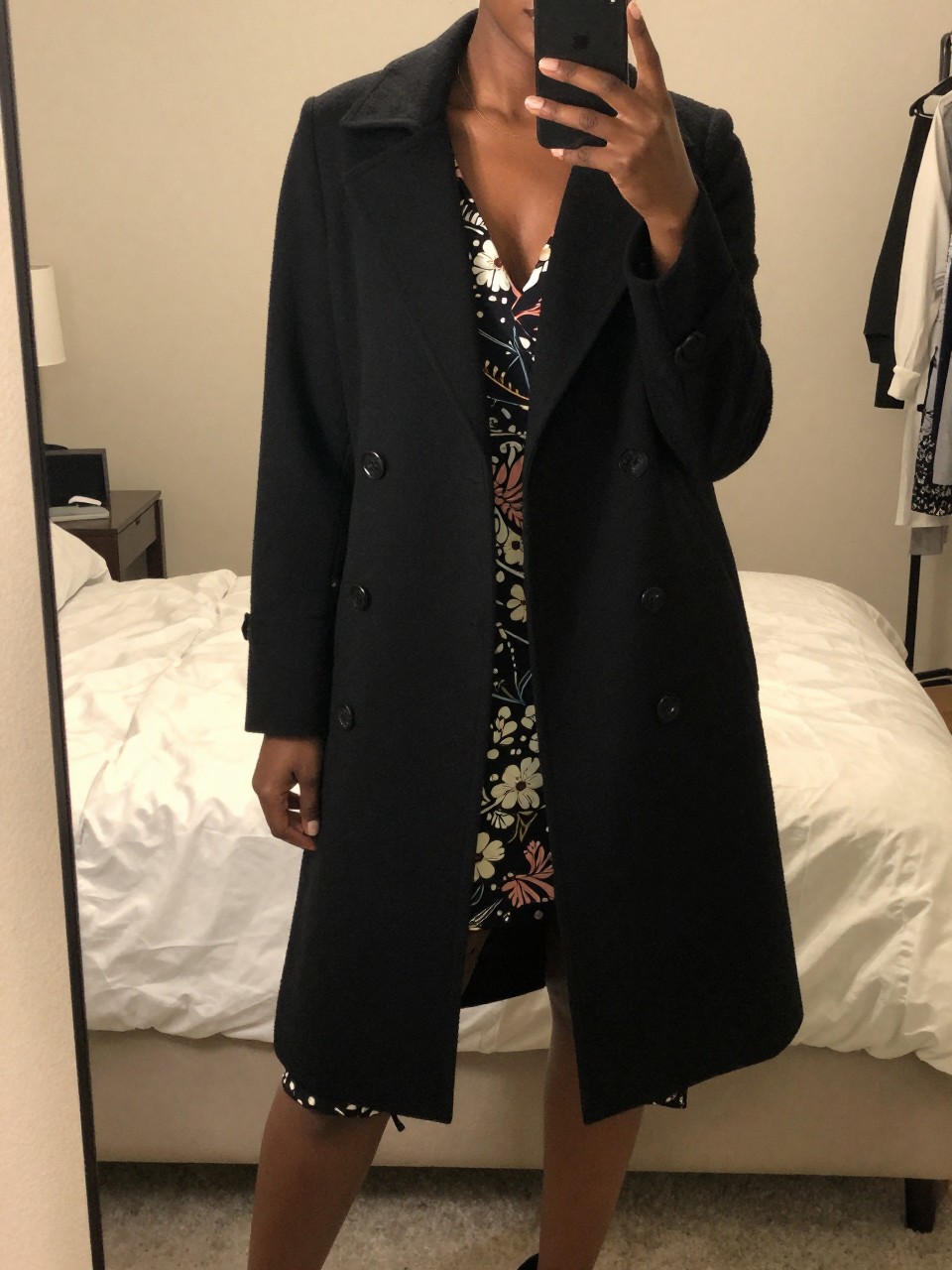 iPhone mirror selfie of a Black woman wearing a black swing coat over a printed dress, standing with one hand holding her phone at her side, cropped at NECK so face doesn't show, in a feminine-themed bedroom, warm moody lighting, iPhone photo quality.