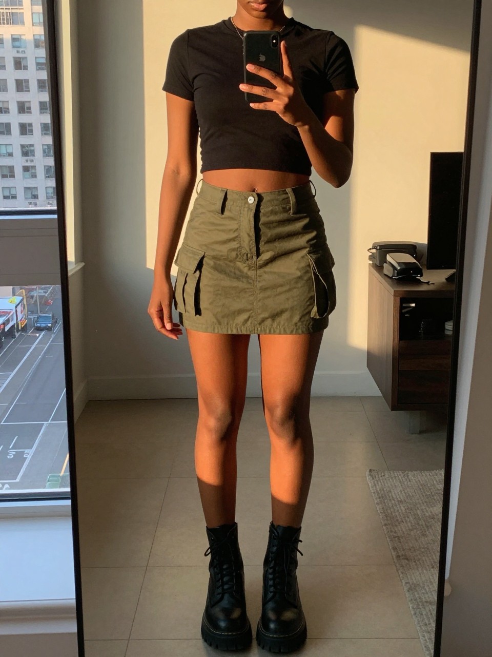 iPhone mirror selfie of a black woman wearing a cropped black band tee with an olive green utility mini skirt and chunky combat boots, one hand holding her phone at chest level, cropped at NECK so face doesn't show, in a city apartment, evening golden hour, iPhone photo quality.