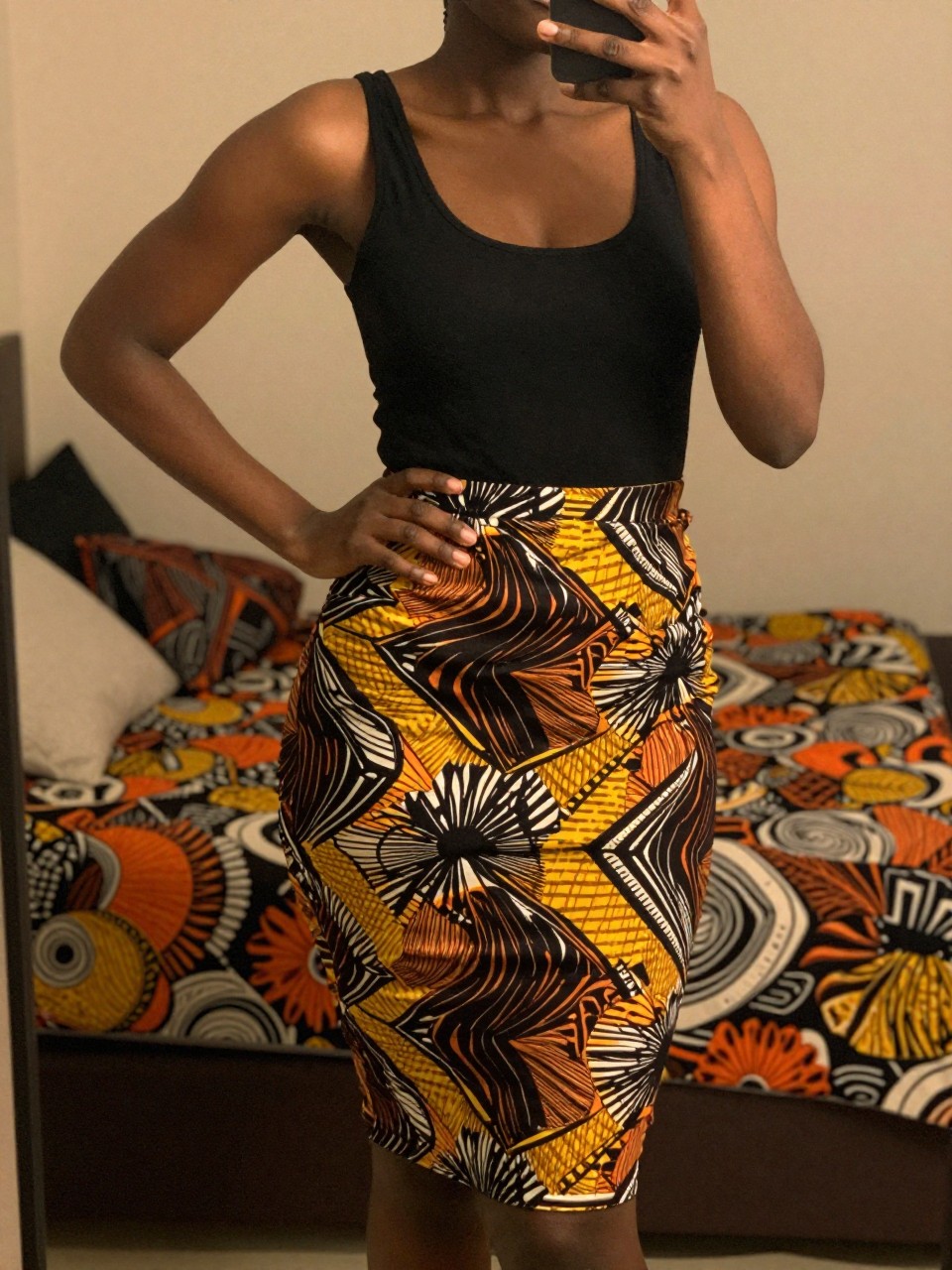 mirror selfie of a black woman in an Ankara print skirt and black tank top, one hand on hip, cropped at NECK so face doesn't show, in a feminine-themed bedroom with warm moody lighting, iPhone photo quality.