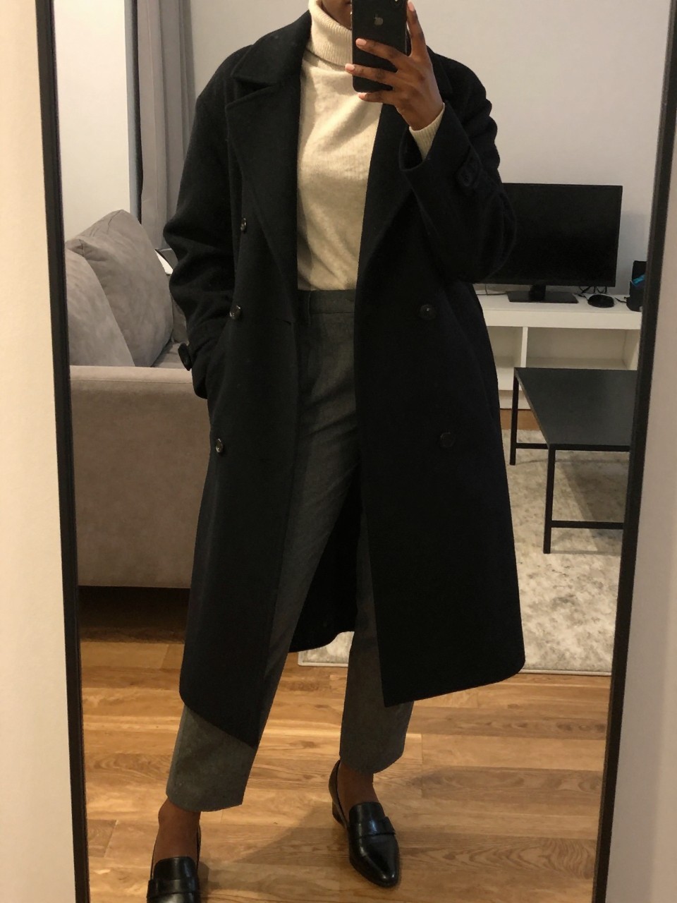 iPhone mirror selfie of a Black woman wearing a long black wool coat over a cream turtleneck and charcoal trousers, standing with her weight on one leg and hands casually in her coat pockets, cropped at NECK so face doesn't show, in a city apartment, warm moody lighting, iPhone photo quality.