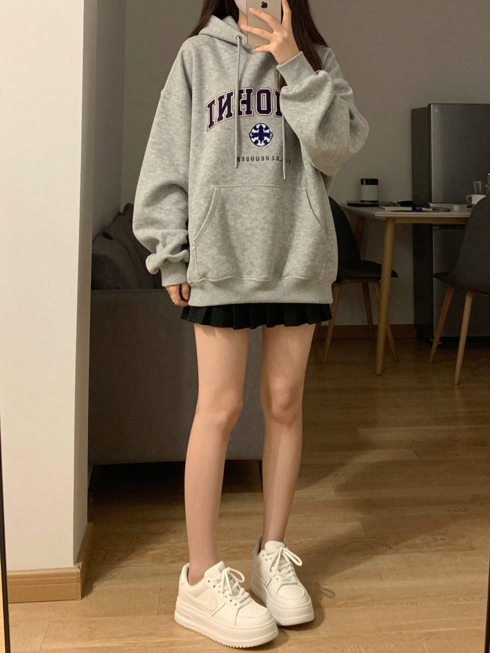 iPhone mirror selfie of a woman wearing an oversized gray graphic hoodie with a black pleated tennis skirt and chunky white platform sneakers, one hand holding her phone at her side, cropped at NECK so face doesn't show, in a city apartment, warm moody lighting, iPhone photo quality.