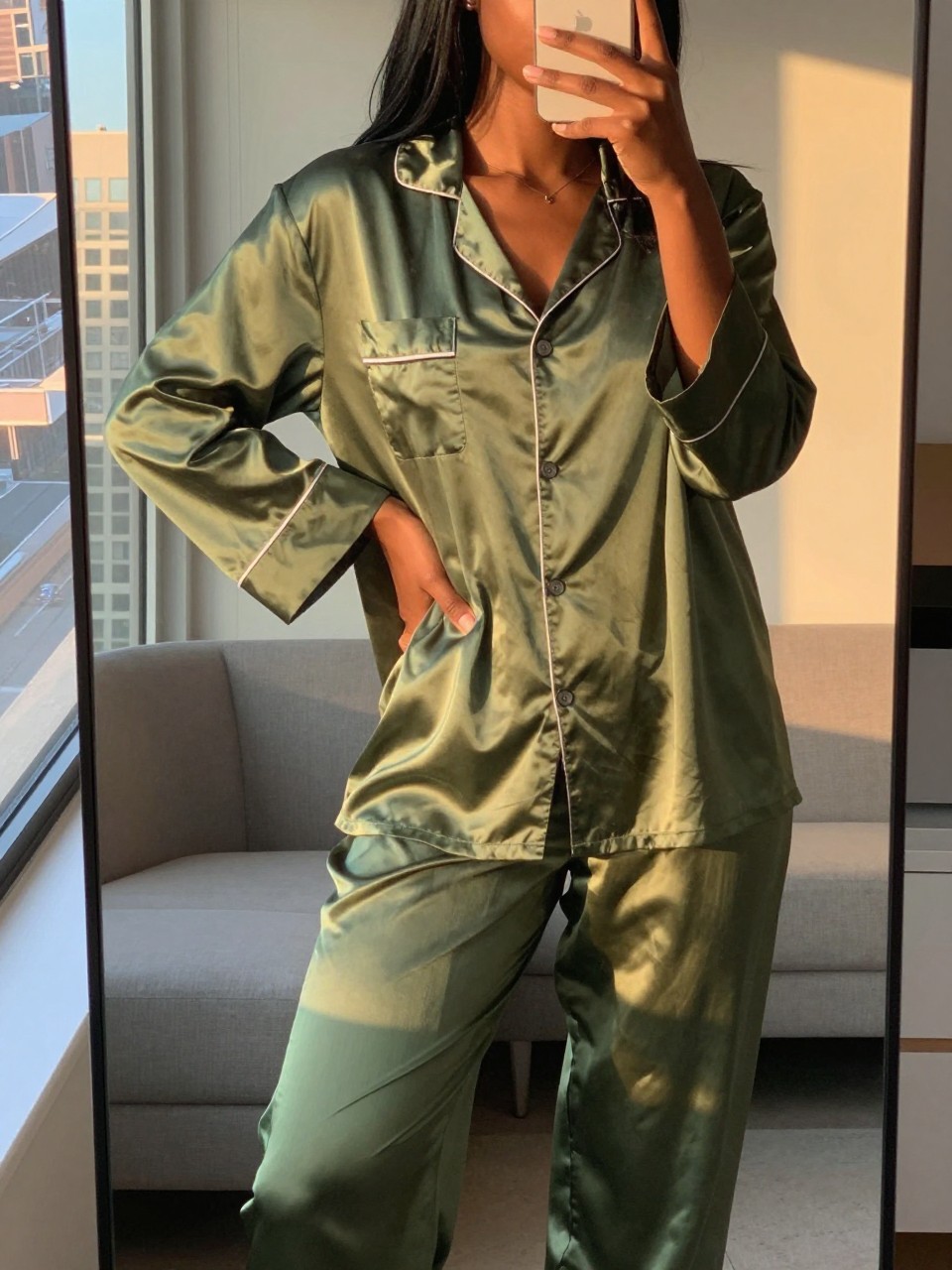 iPhone mirror selfie of a black woman in an olive green silk pajama set and robe, relaxed pose with hand on hip, cropped at NECK so face doesn't show, in a city apartment during evening golden hour, iPhone photo quality.