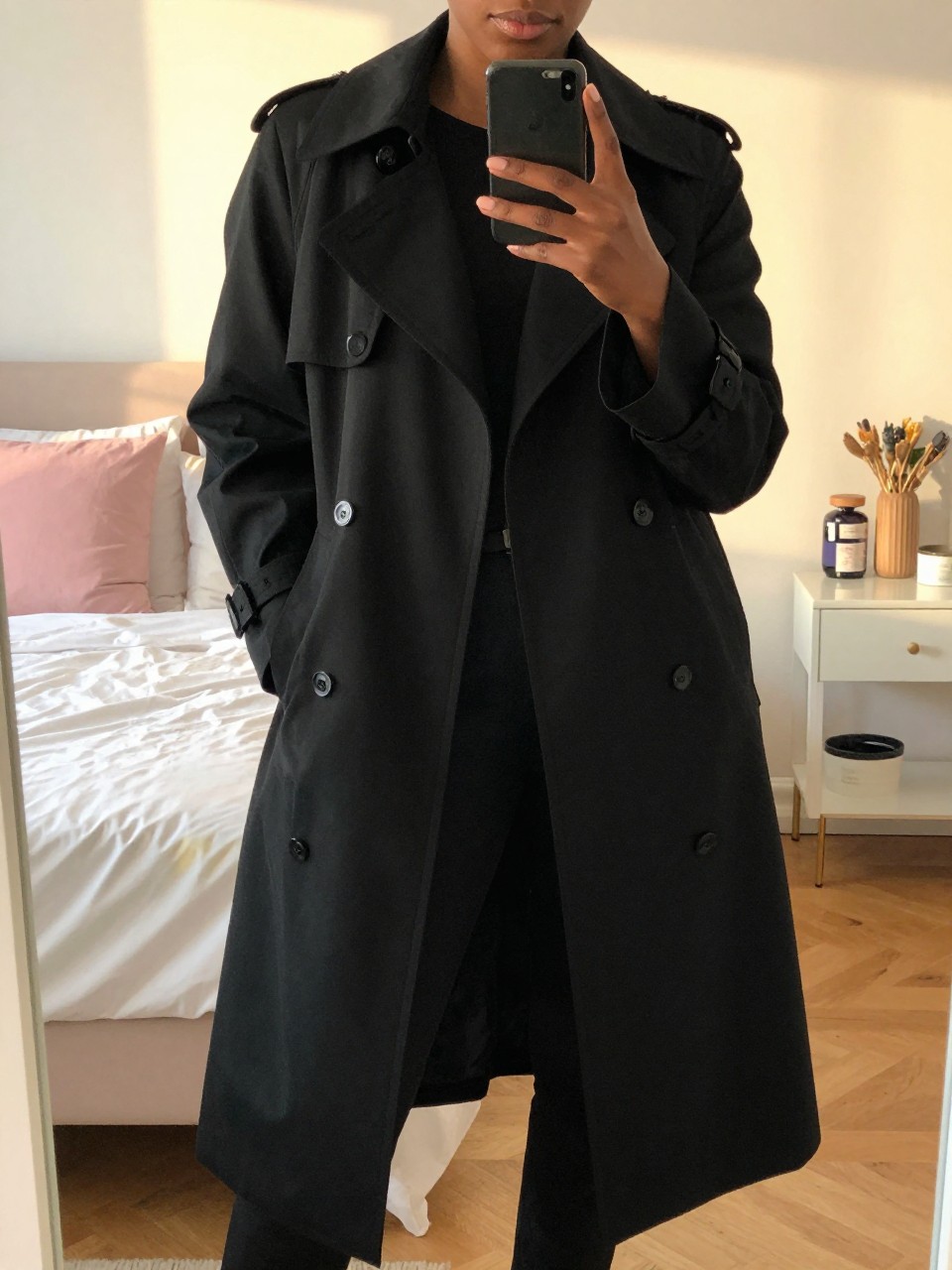 iPhone mirror selfie of a Black woman wearing a black trench coat over an all-black outfit, posed with one hand holding her phone and the other in her pocket, cropped at NECK so face doesn't show, in a bright feminine-themed bedroom, evening golden hour light, iPhone photo quality.