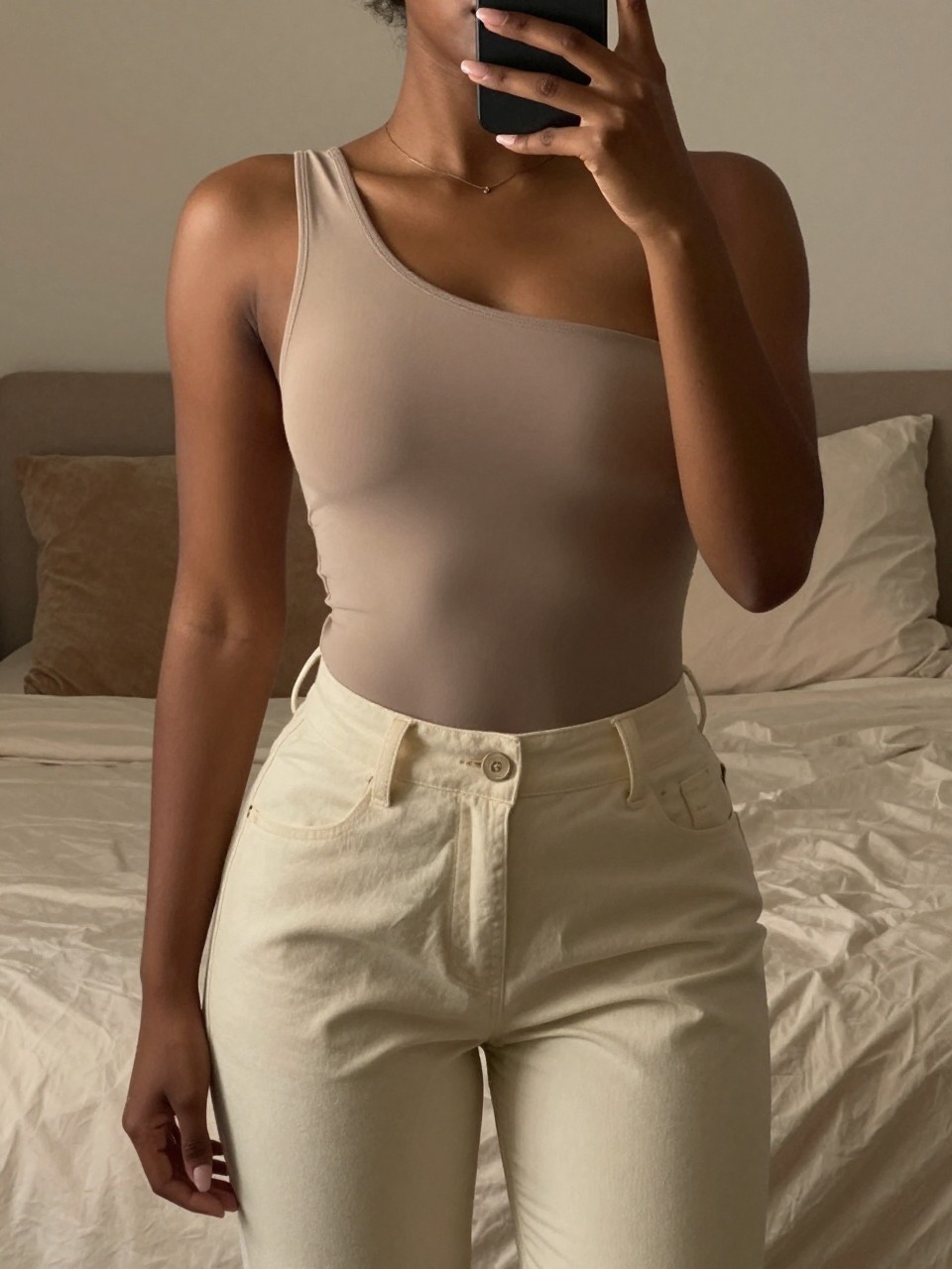 iPhone mirror selfie of a black woman in a one-shoulder bodysuit and cream trousers, posed with arm slightly bent, cropped at NECK so face doesn't show, in a feminine-themed bedroom with warm moody lighting, iPhone photo quality.