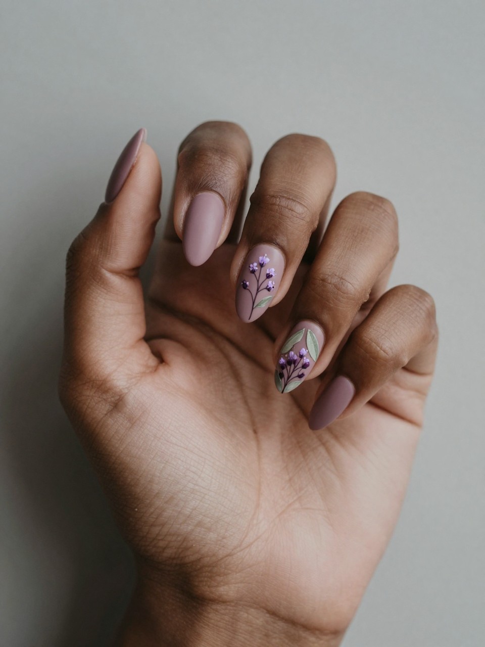 Bird's-eye view of black feminine hand with almond. The nails are a matte mauve. Delicate floral sprigs with tiny purple buds and sage green leaves are painted on each nail. The mood is rustic and serene.
