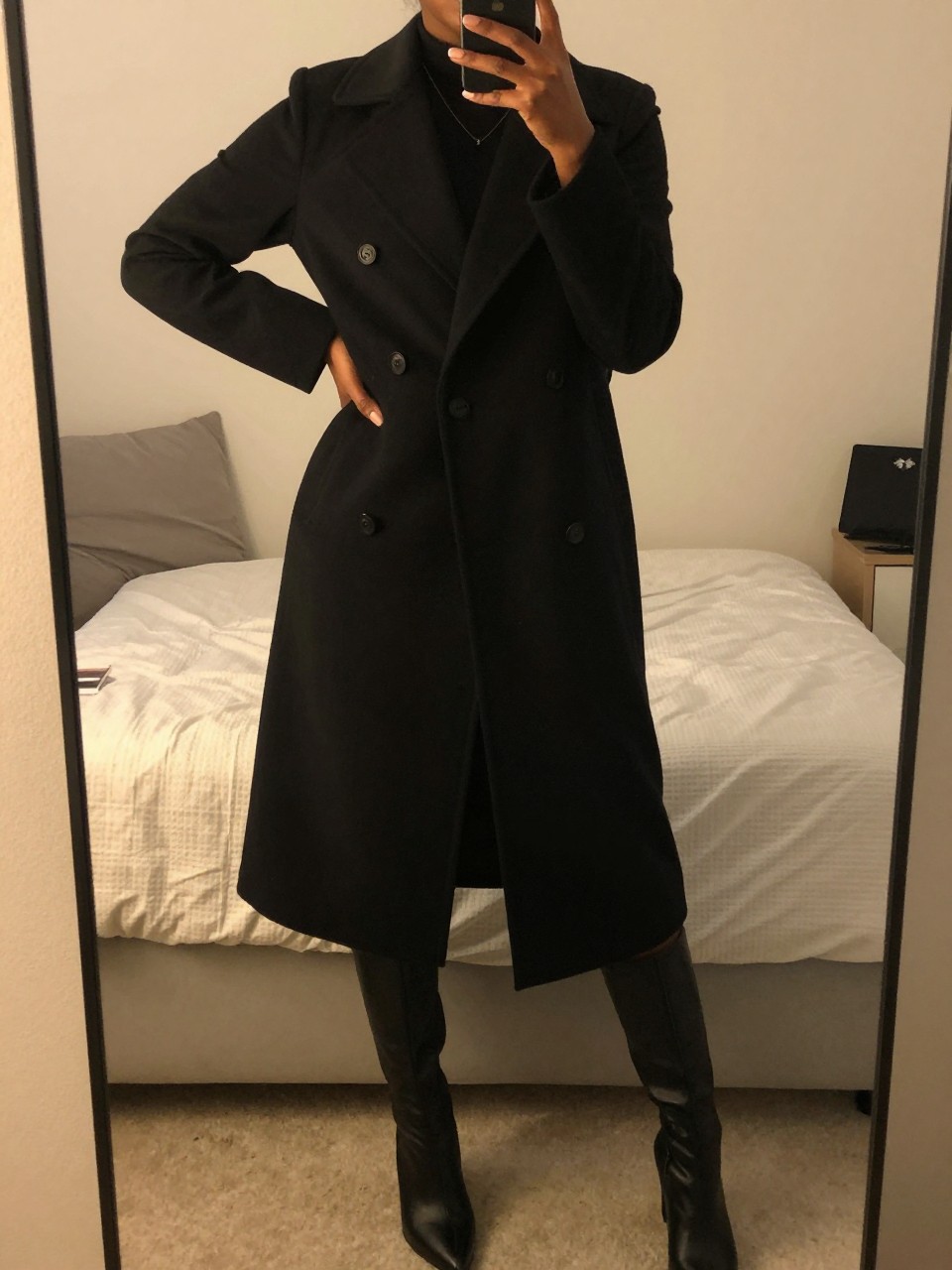 iPhone mirror selfie of a Black woman wearing a double-breasted black coat over a midi skirt and knee-high boots, standing with one hand on her hip, cropped at NECK so face doesn't show, in a feminine-themed bedroom, evening golden hour light, iPhone photo quality.