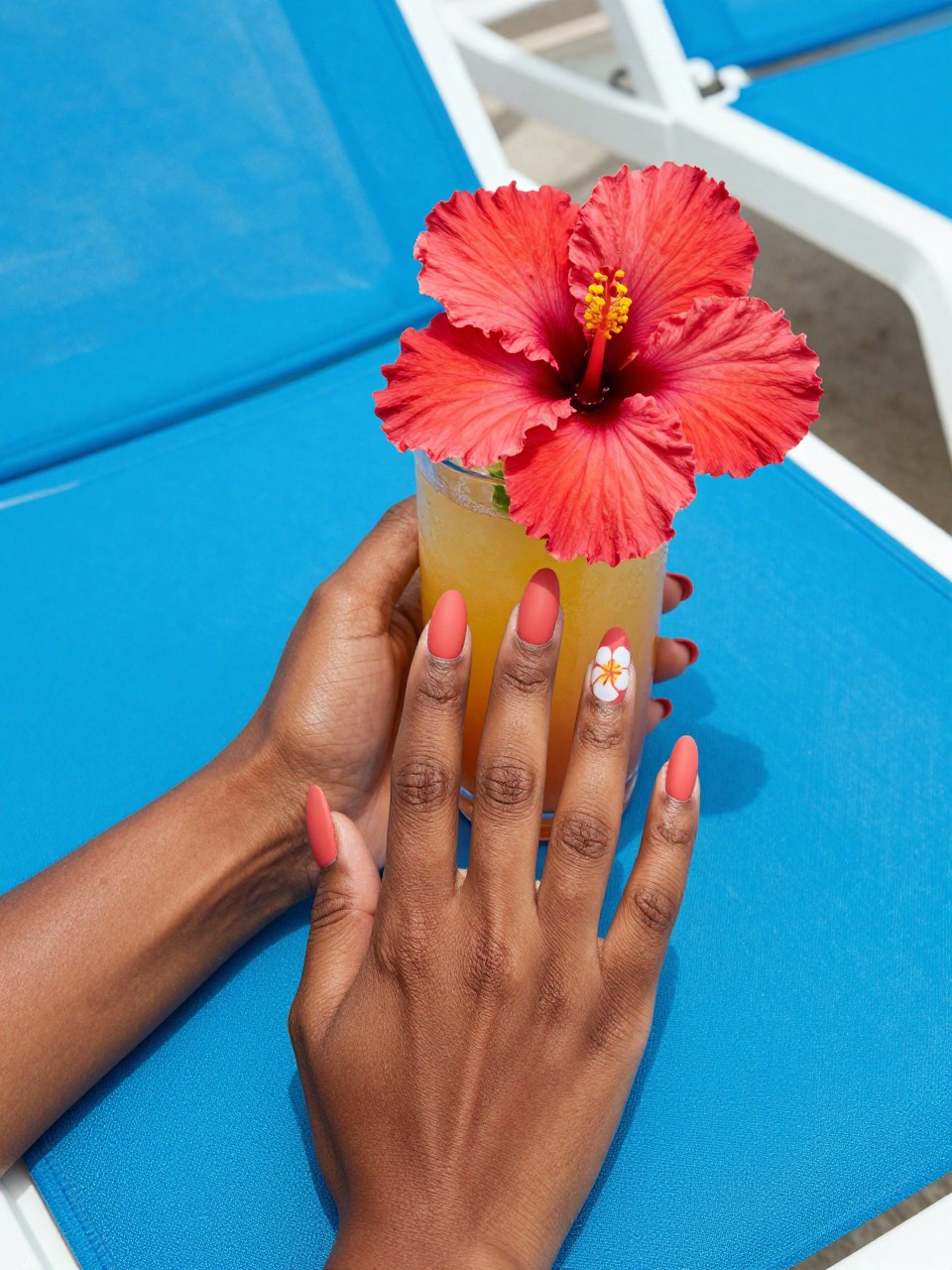 Bird's-eye view of black woman hands with almond nails next to a tropical drink adorned with a hibiscus flower. The nails are a matte coral. One accent nail features a white hibiscus with a yellow center and a small green leaf. The background is a bright blue poolside lounge chair.