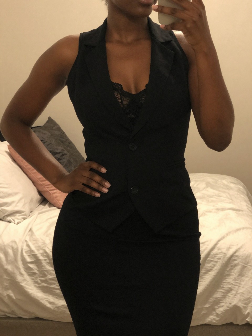 iPhone mirror selfie of a black woman wearing a black vest over a lace camisole, standing with one hand on her hip, cropped at NECK so face doesn't show, in a feminine-themed bedroom, warm moody lighting, iPhone photo quality.