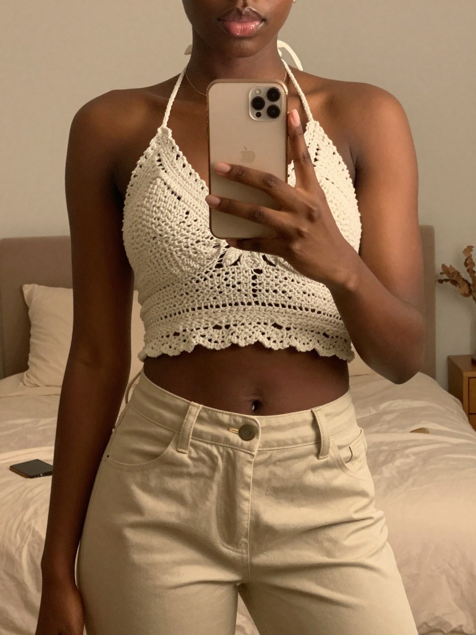 iPhone mirror selfie of a black woman in a crochet halter top and beige pants, holding phone slightly low, cropped at NECK so face doesn't show, in a feminine-themed bedroom with warm moody lighting, iPhone photo quality.