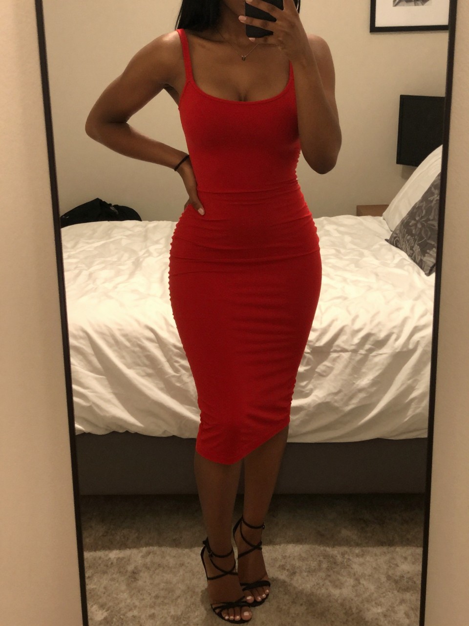 iPhone mirror selfie of a black woman in a red bodycon dress and strappy heels, angled slightly with hand on hip, cropped at NECK so face doesn't show, in a feminine-themed bedroom with warm moody lighting, iPhone photo quality.