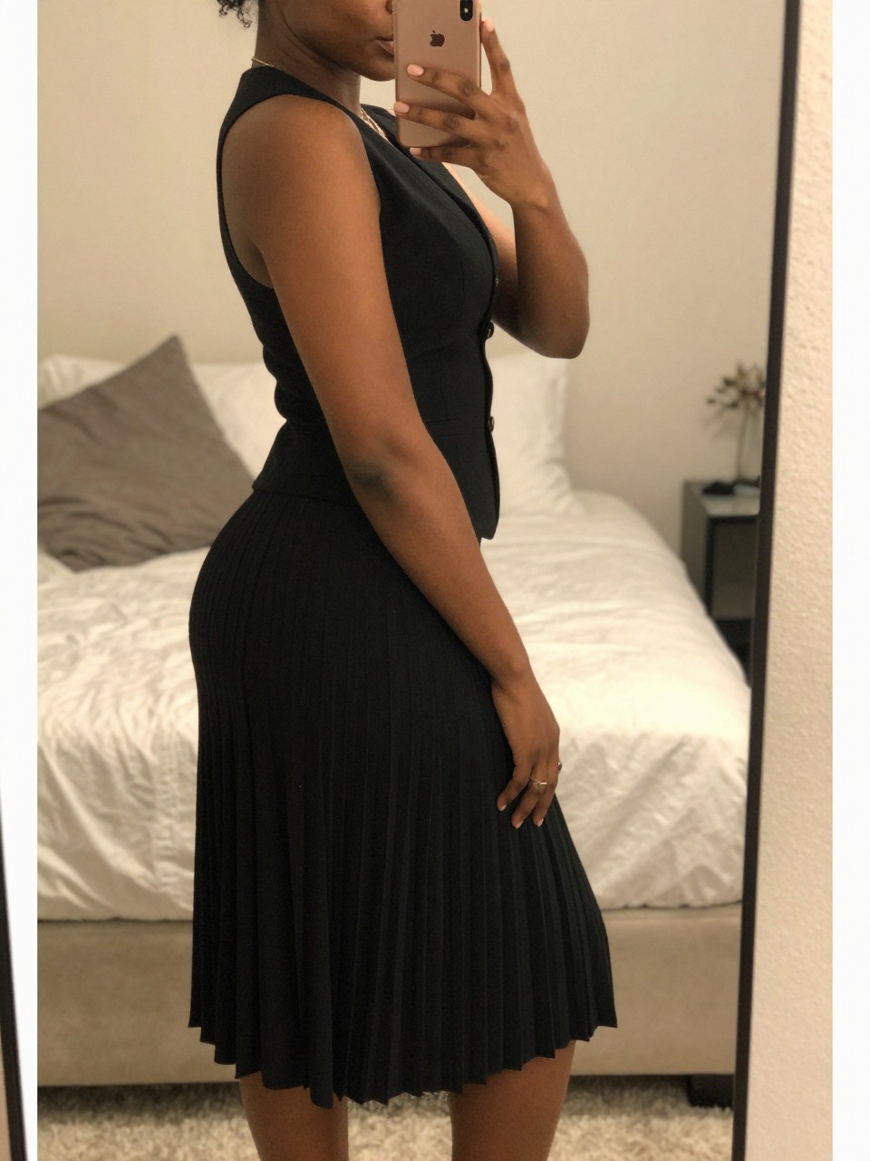 iPhone mirror selfie of a black woman wearing a black vest with a pleated midi skirt, posed with her phone in front and weight back, cropped at NECK so face doesn't show, in a feminine-themed bedroom, warm moody lighting, iPhone photo quality.
