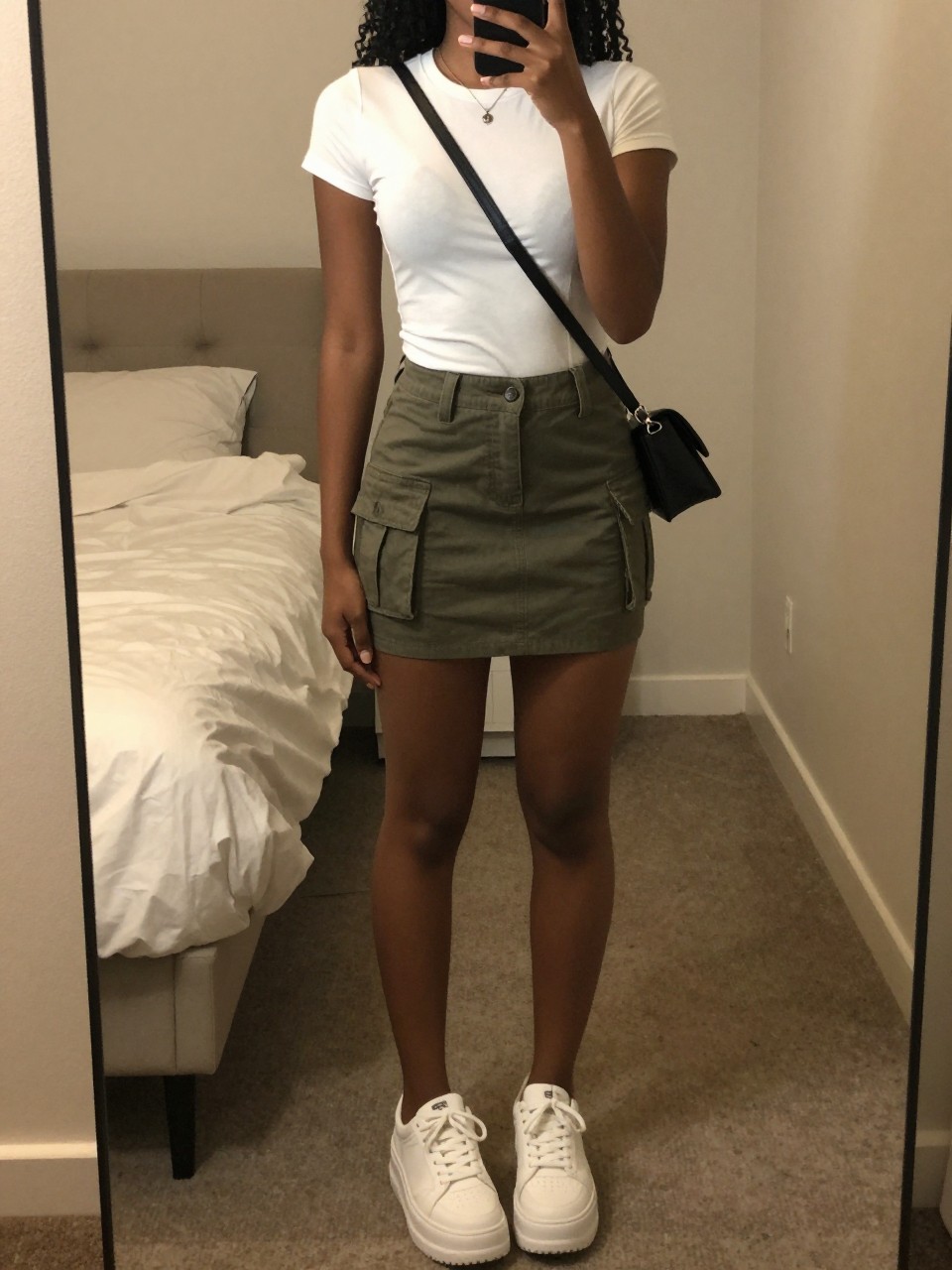 iPhone mirror selfie of a black woman wearing a fitted white t-shirt tucked into an olive green cargo mini skirt with chunky white platform sneakers and a small black crossbody bag, standing in a relaxed pose with one hand in the skirt pocket, cropped at NECK so face doesn't show, in a feminine-themed bedroom, warm moody lighting, iPhone photo quality.