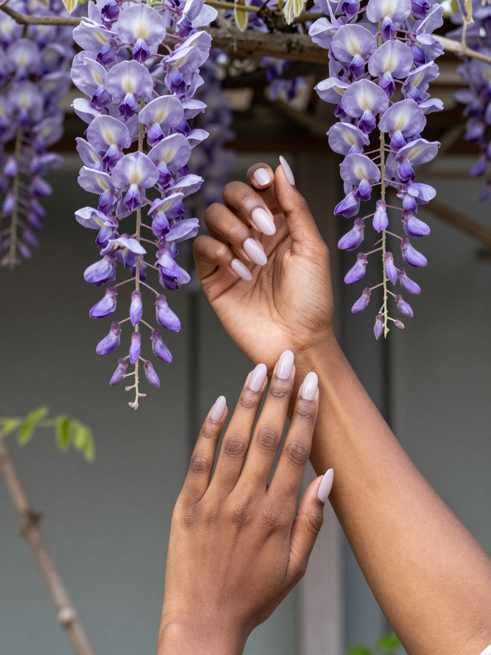 Bird's-eye view of black feminine hands with long, oval nails posed near a blooming wisteria trellis. The nails show a soft lilac-to-white ombré. A thin vine with hanging clusters of tiny purple wisteria flowers trails down each nail. The light is dappled and hazy.