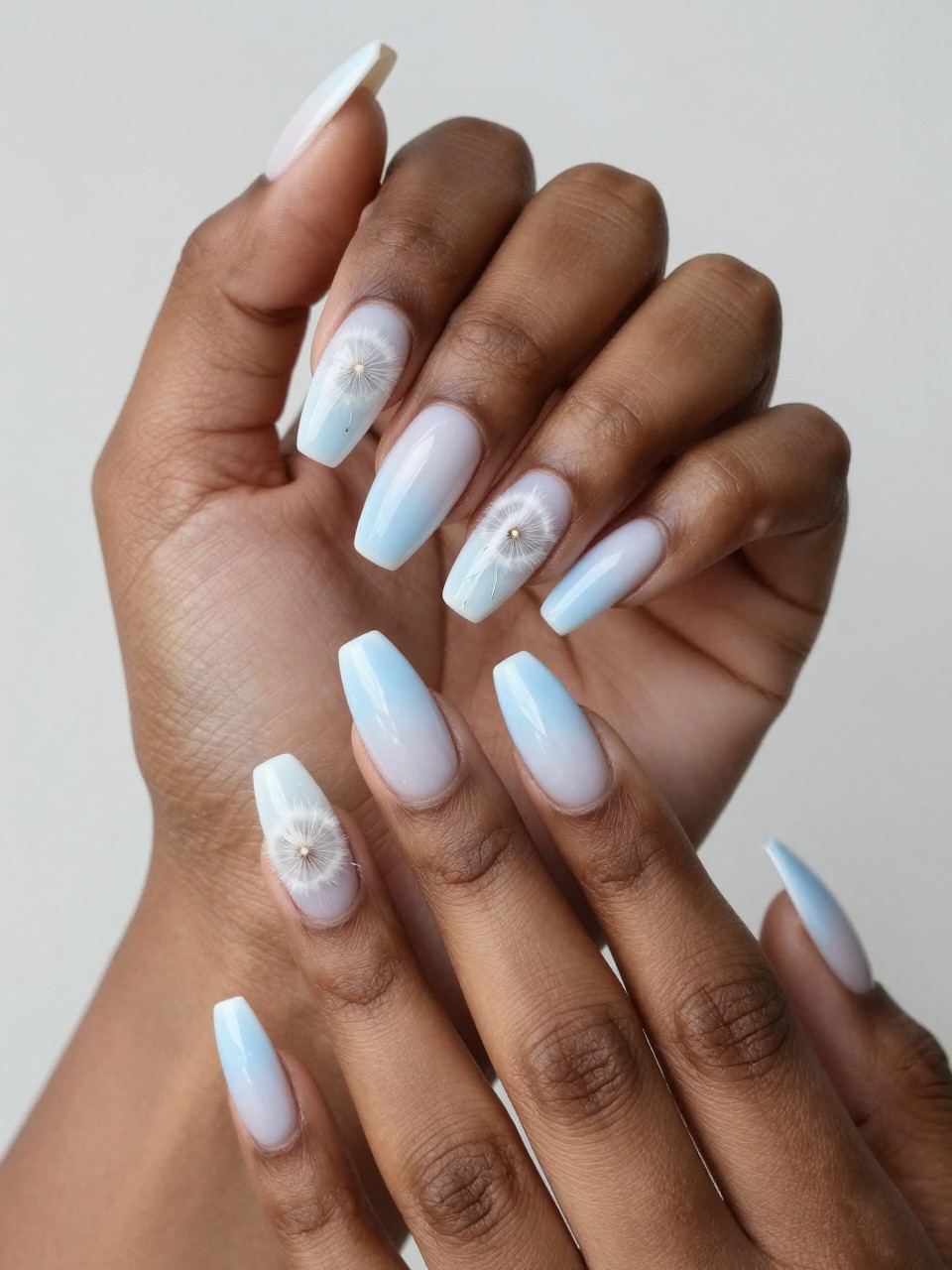 Angled view of black feminine hands with long nails. The nails show a gentle white-to-light-blue ombré. A delicate dandelion puff is painted on each nail, with a few white seed lines drifting off the tip. The effect is soft, poetic, and airy.