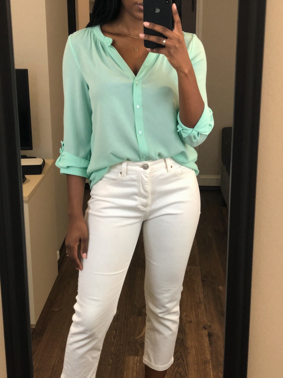 iPhone mirror selfie of a Black woman wearing a seafoam green blouse with white cropped pants, standing casually with her phone at her side, cropped at NECK so face doesn't show, in a city apartment, warm moody lighting, iPhone photo quality.