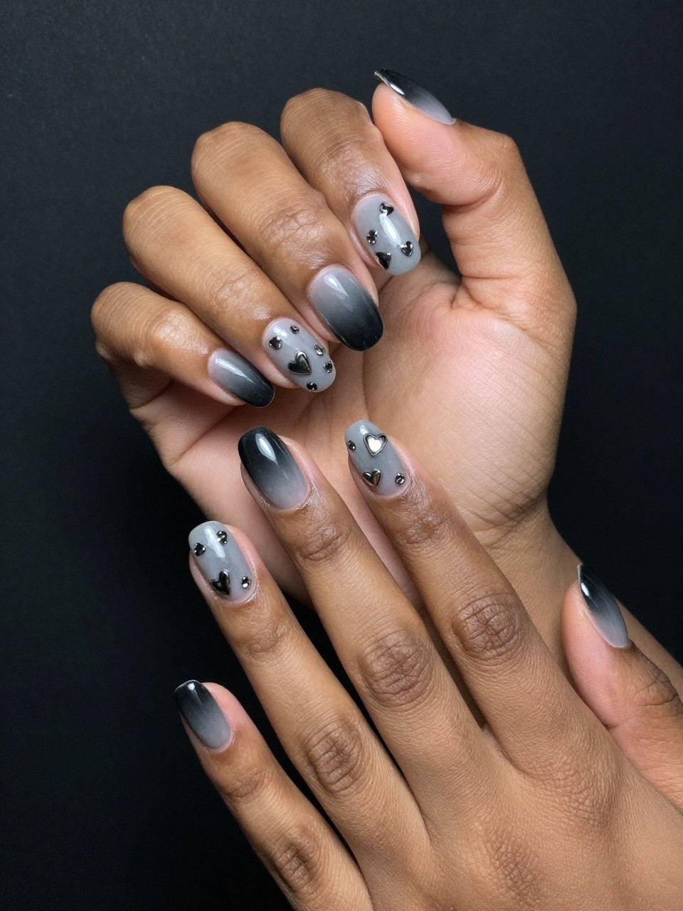 Angled view of black feminine hands with oval nails against a dark, moody backdrop. The nails show a smoky grey-to-black ombré. Various small, reflective chrome hearts are scattered across each nail. The lighting is soft and dramatic, with the hearts catching occasional gleams.