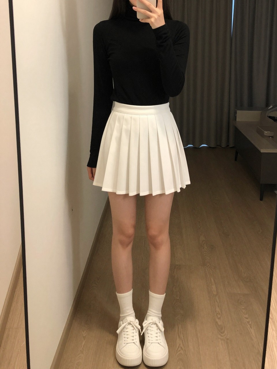 iPhone mirror selfie of a woman wearing a black turtleneck tucked into a white pleated tennis skirt with white platform sneakers and crew socks, one hand holding her phone at her side, cropped at NECK so face doesn't show, in a city apartment, warm moody lighting, iPhone photo quality.