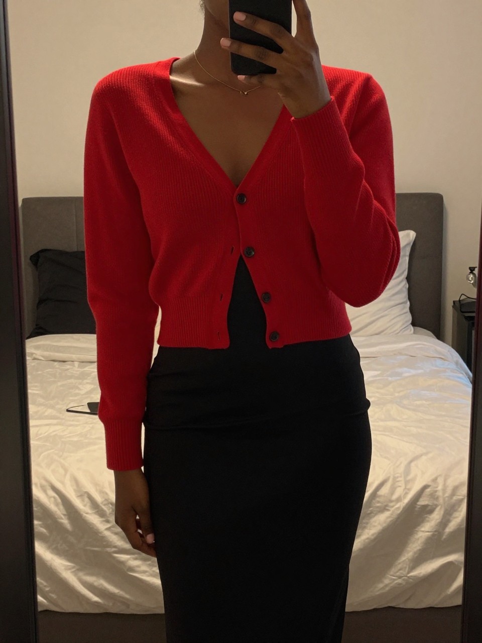 iPhone mirror selfie of a black woman in a red cardigan and black slip skirt, relaxed pose, cropped at NECK so face doesn't show, in a feminine-themed bedroom with warm moody lighting, iPhone photo quality.