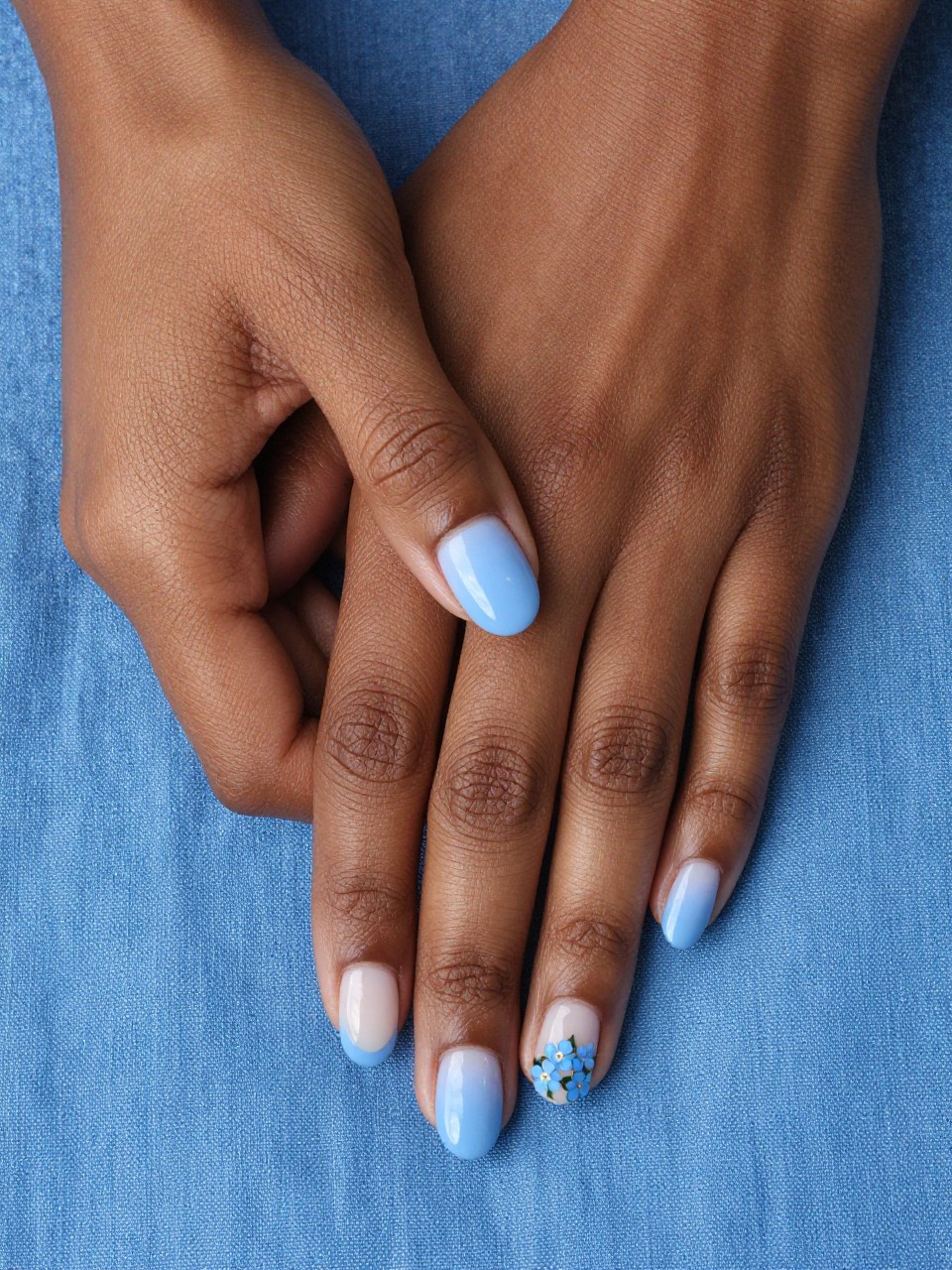 Overhead shot of black feminine hands with oval nails resting on a blue linen cloth. The French tips are a powdery sky blue. On the natural nail base, a few tiny blue forget-me-not flowers are painted near the cuticles. The mood is serene and pretty.