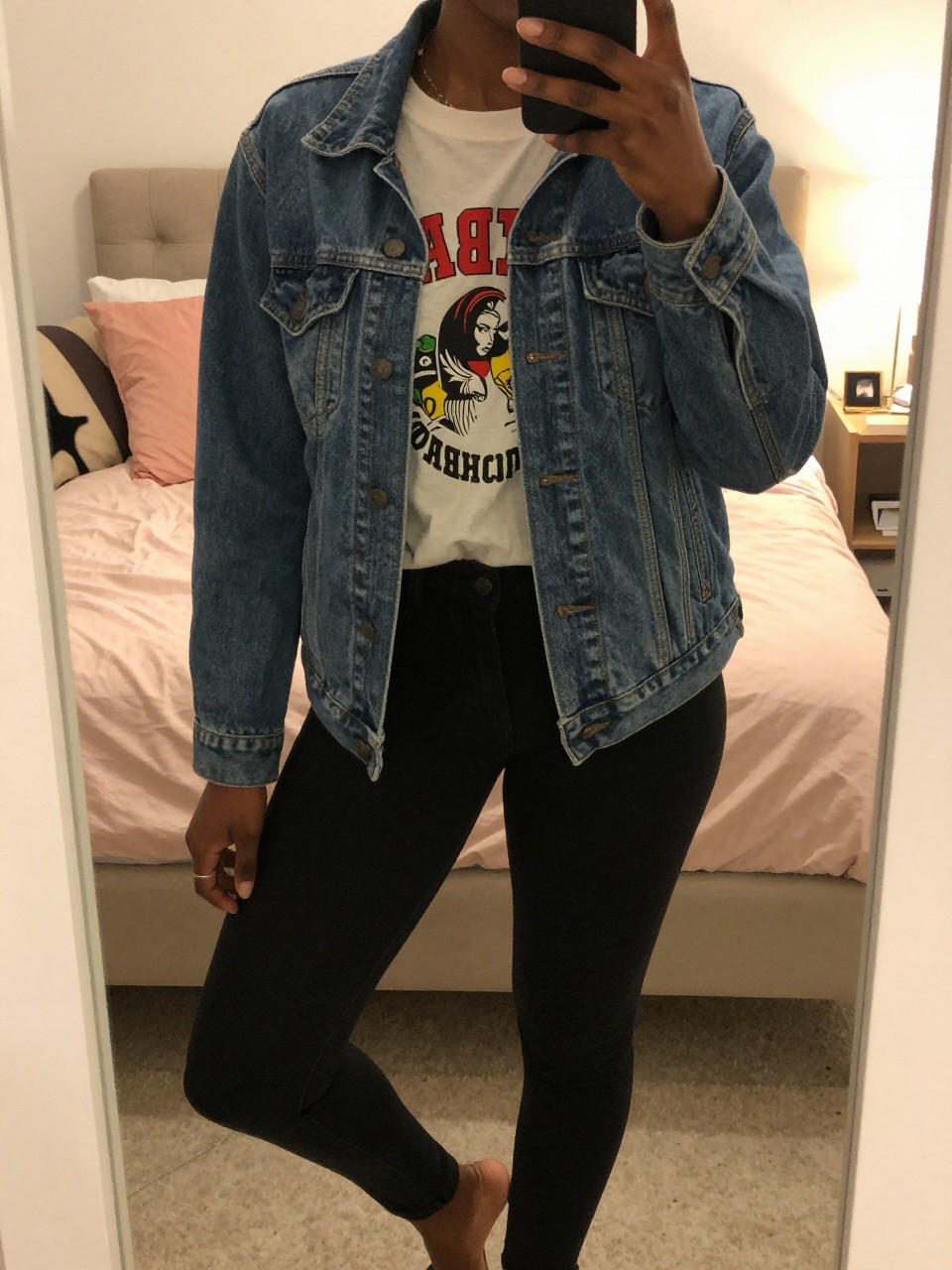 mirror selfie of a black woman in a jean jacket and graphic tee, relaxed pose with one foot forward, cropped at NECK so face doesn't show, in a feminine-themed bedroom with warm moody lighting, iPhone photo quality.