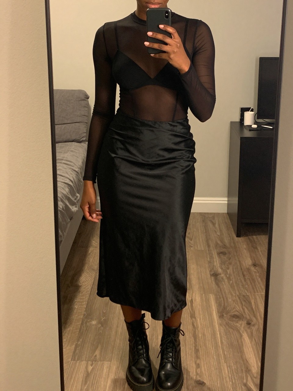 iPhone mirror selfie of a black woman wearing a black sheer mesh top over a bralette with a black satin slip midi skirt and black combat boots, one hand holding her phone at chest level, cropped at NECK so face doesn't show, in a city apartment, warm moody lighting, iPhone photo quality.