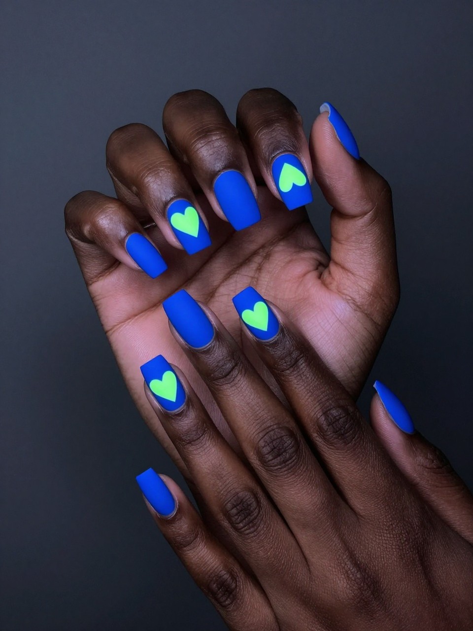 Feminine black hands in a dimly lit setting with a subtle UV light source. The nails are matte electric blue. Neon green hearts appear to glow on each nail. The look is modern, playful, and perfect for a night out.