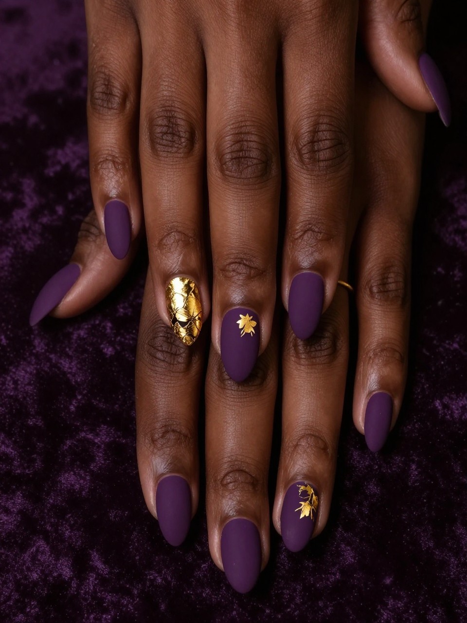Close-up, top-down view of black feminine hands with almond nails against a dark velvet background. The nails are a flat matte plum. On the ring fingers, fragile gold leaf is arranged in a crumbling, floral-inspired pattern. Warm, dim lighting makes the gold gleam against the deep purple.