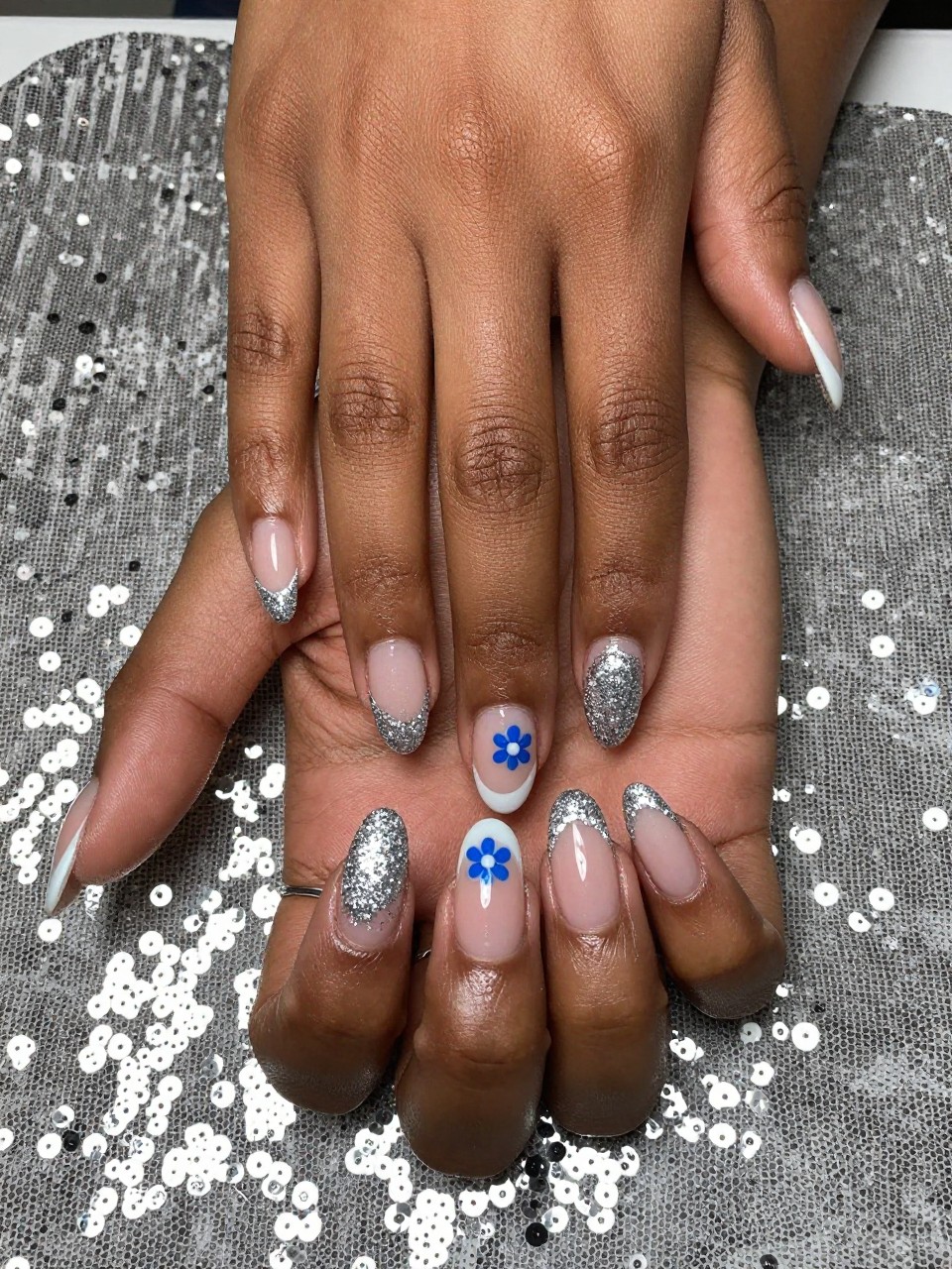Top-down view of black feminine hands with coffin nails resting on a sequined cloth. The French tips are made of fine silver glitter. On the natural nail bed, just below the glitter line, a tiny blue flower with a white center is painted on each nail. The look is glamorous and detailed.