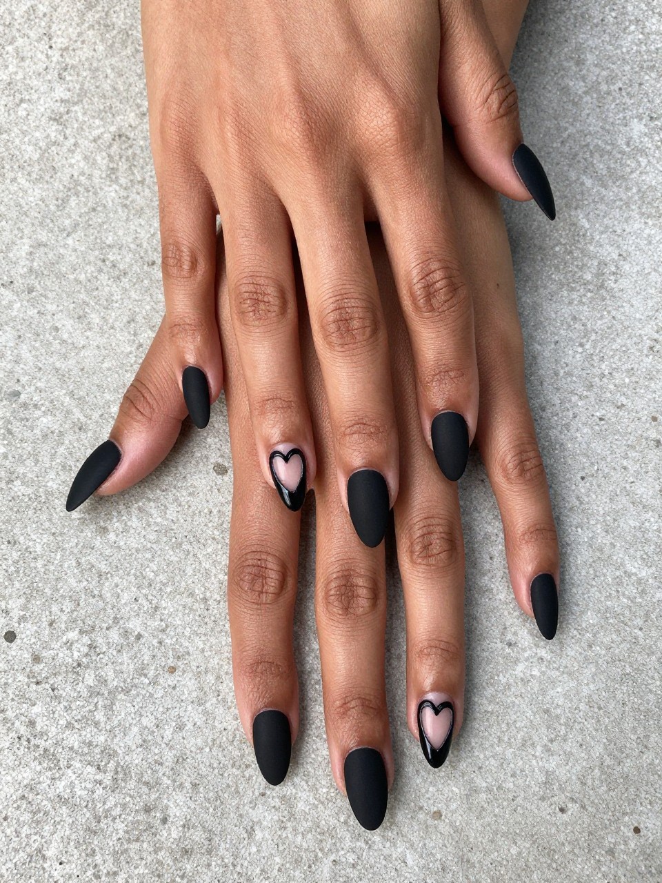 Overhead, top-down view of black feminine hands with long, coffin-shaped nails resting on a rough concrete surface. The nails are a perfect matte black. A single, glossy black heart outline is centered on each nail, catching the light sharply against the dull base. The mood is urban and sophisticated.