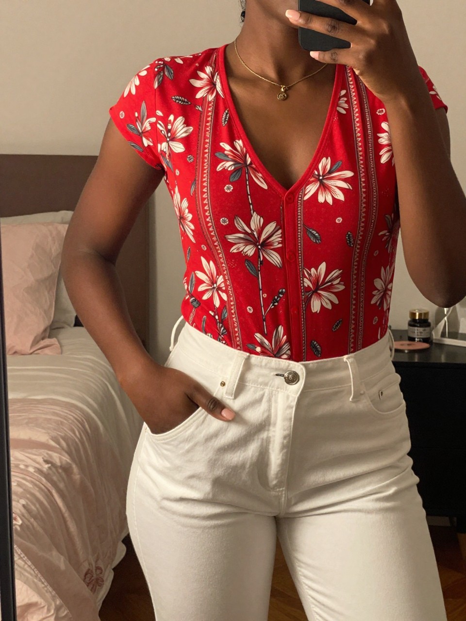 mirror selfie of a black woman in a red print top and white pants, hand in pocket, cropped at NECK so face doesn't show, in a feminine-themed bedroom with warm moody lighting, iPhone photo quality.