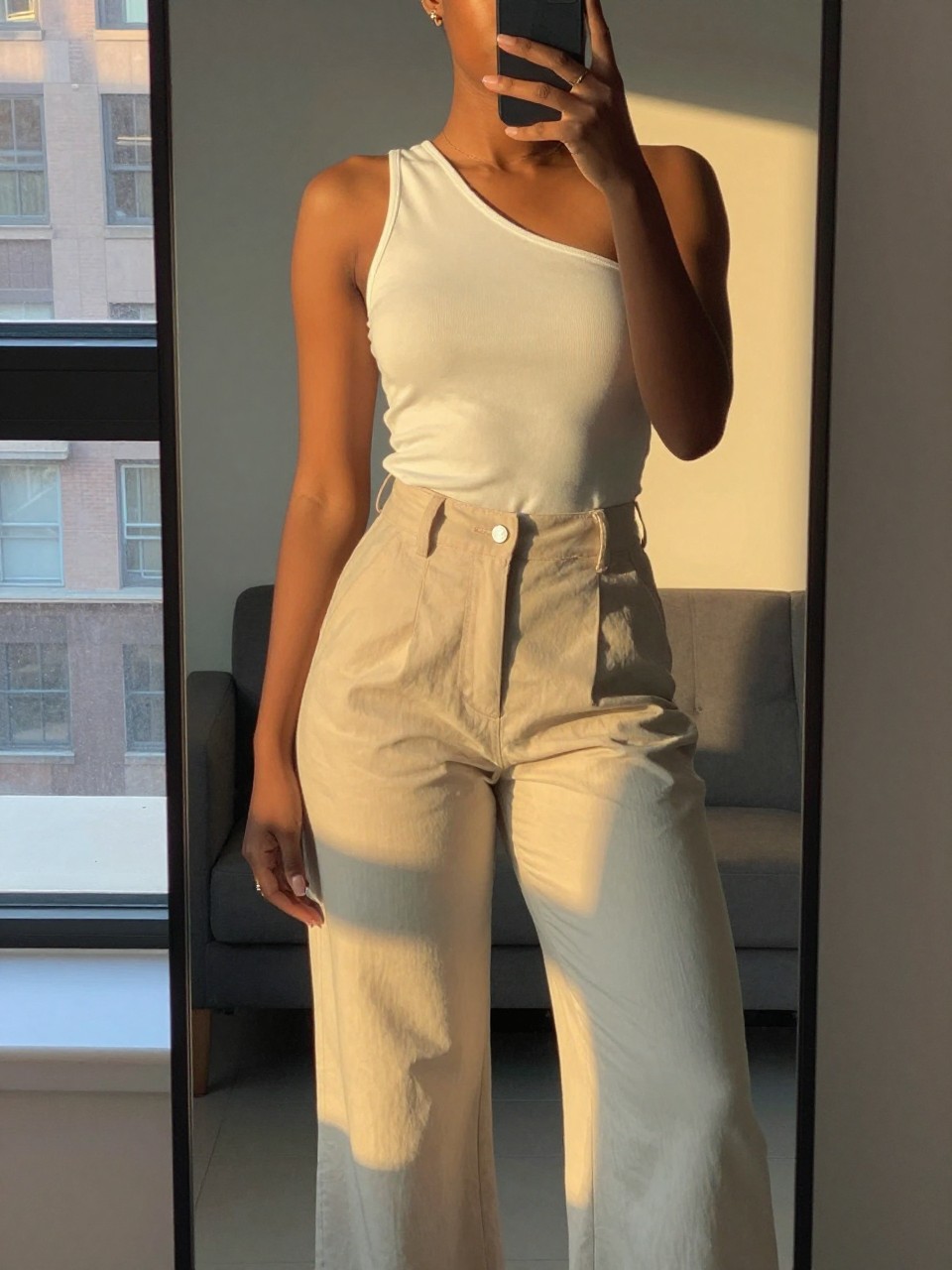 iPhone mirror selfie of a black woman in a one-shoulder top and palazzo pants, one hand behind head, cropped at NECK so face doesn't show, in a city apartment during evening golden hour, iPhone photo quality.