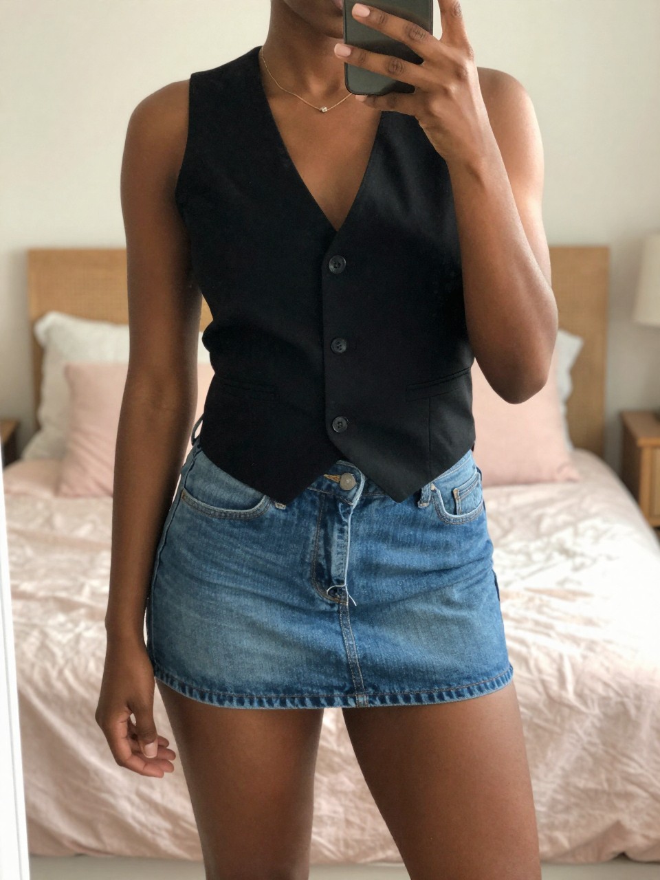 iPhone mirror selfie of a black woman wearing a black vest with a denim mini skirt, standing with her weight shifted to one side, cropped at NECK so face doesn't show, in a bright feminine-themed bedroom, warm moody lighting, iPhone photo quality.