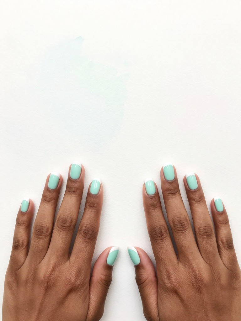 Bird's-eye view of black hands laid flat on a faded pastel watercolor painting. The short, oval nails have a pale seafoam green base with a crisp, white French tip. The colors complement each other softly. The lighting is bright and clear, like an art studio.