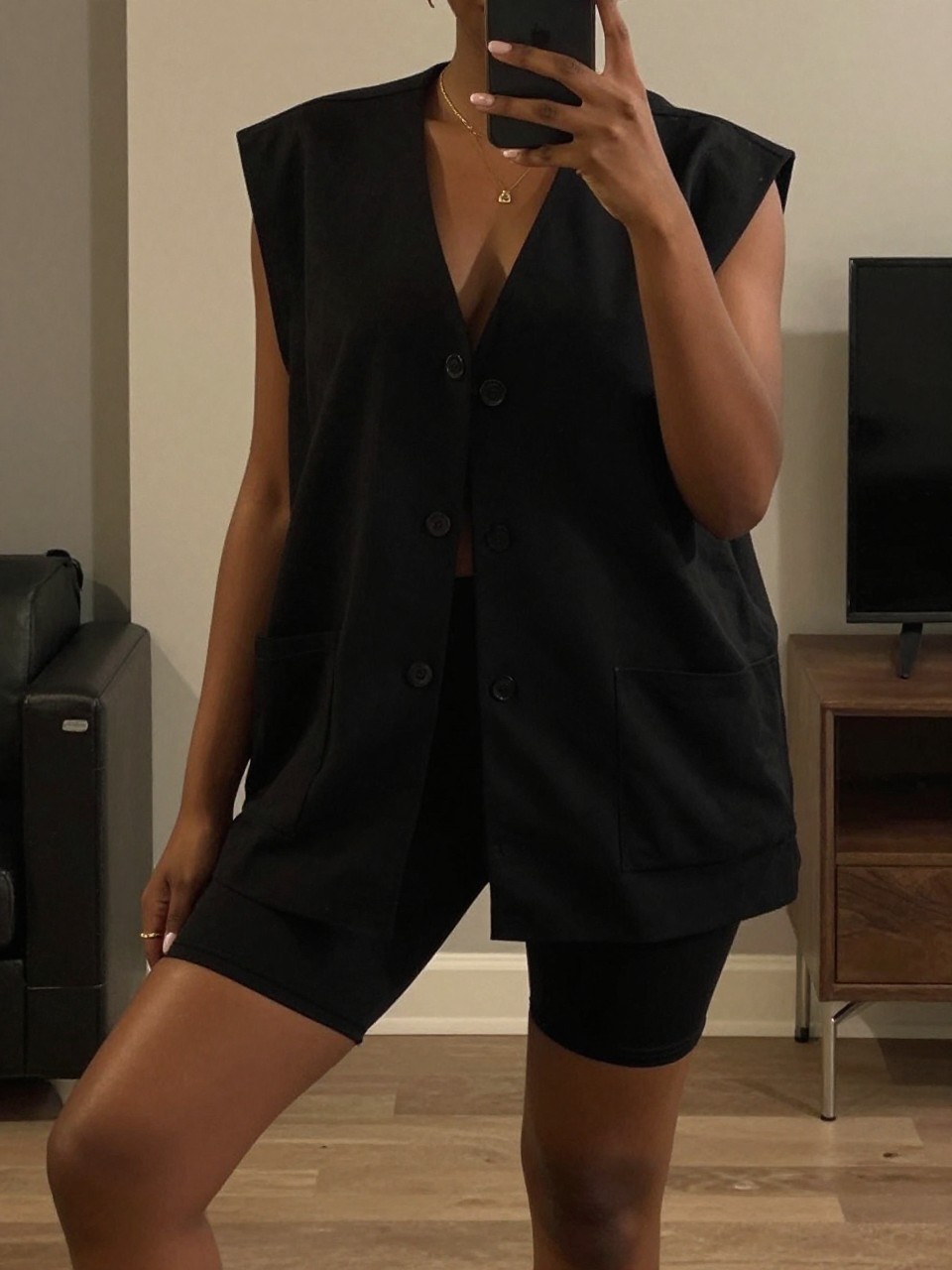 iPhone mirror selfie of a black woman wearing an oversized black vest with biker shorts, posed with her phone low and one knee bent, cropped at NECK so face doesn't show, in a city apartment, warm moody lighting, iPhone photo quality.