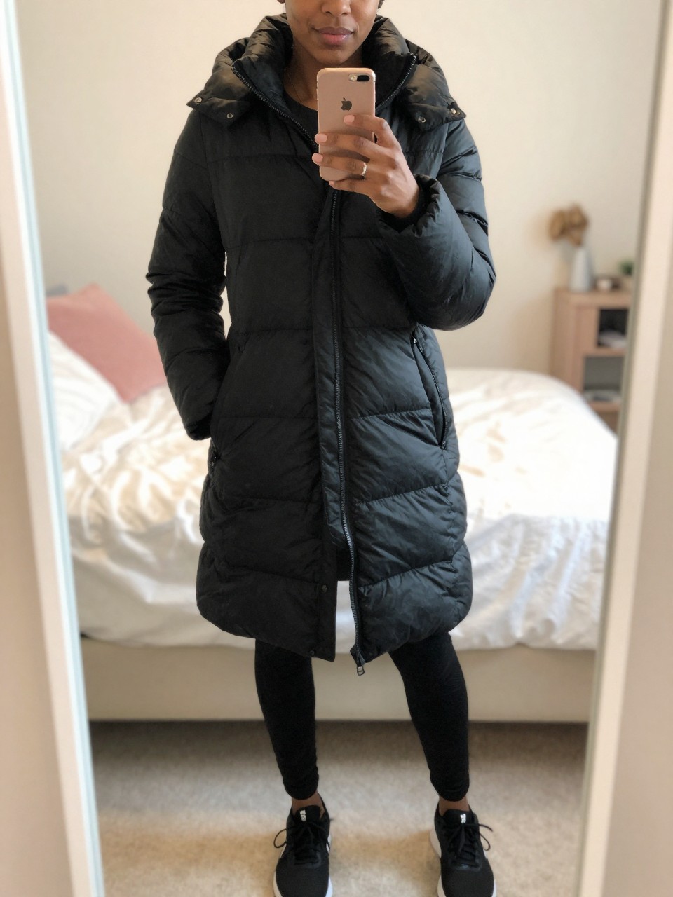 iPhone mirror selfie of a Black woman wearing a sleek black puffer coat with leggings and sneakers, standing with her weight shifted and phone low, cropped at NECK so face doesn't show, in a bright feminine-themed bedroom, warm moody lighting, iPhone photo quality.