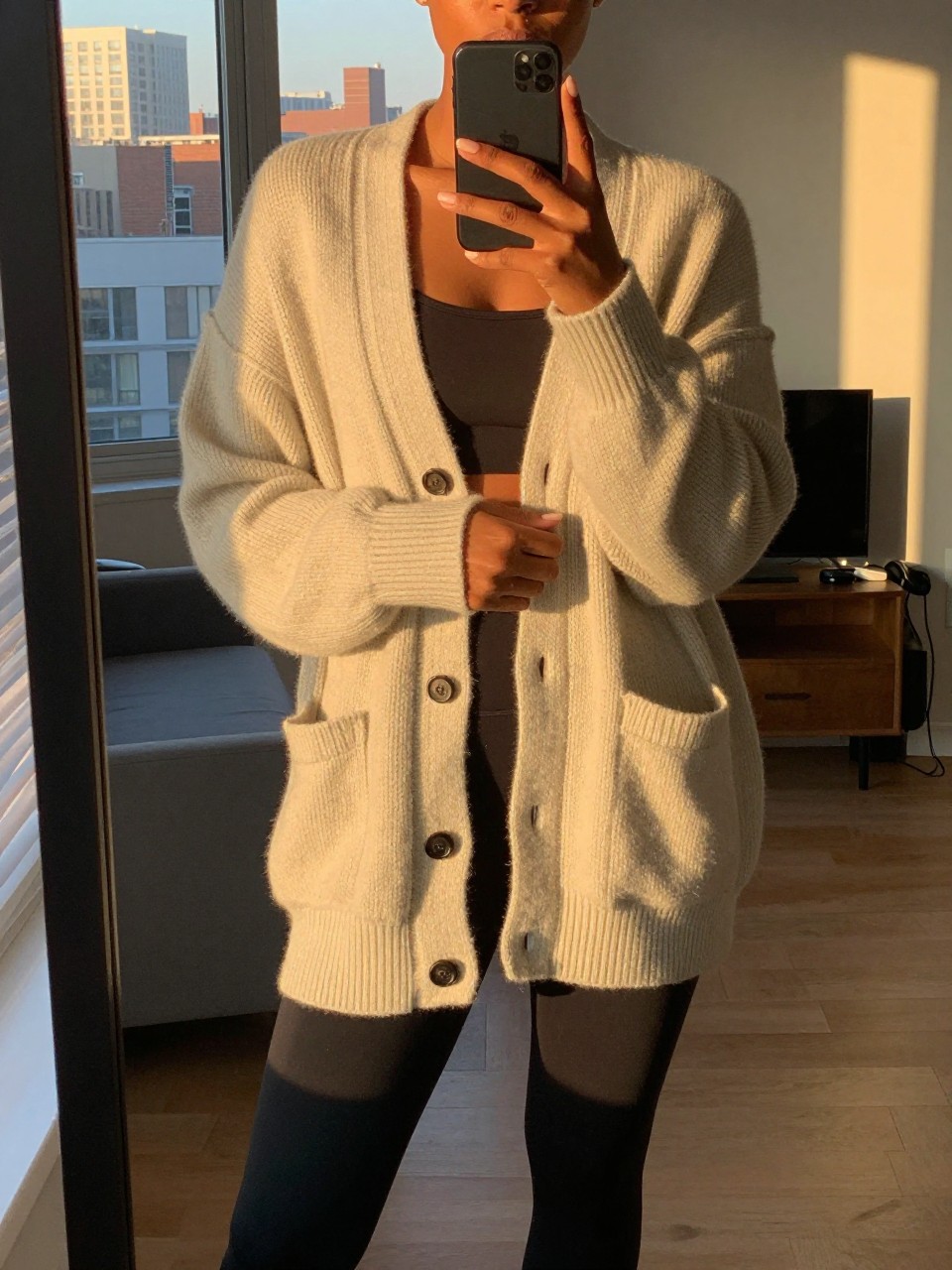 iPhone mirror selfie of a black woman in an oversized cardigan and leggings, wrapped in the cardigan, cropped at NECK so face doesn't show, in a city apartment during evening golden hour, iPhone photo quality.