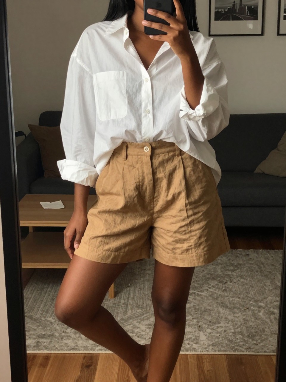 iPhone mirror selfie of a black woman wearing an oversized white button-down and camel linen shorts, standing with her weight on one leg, cropped at NECK so face doesn't show, in a city apartment, warm moody lighting, iPhone photo quality.