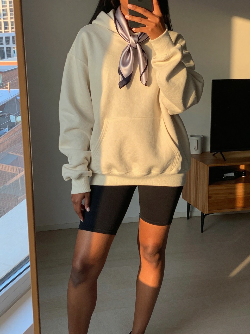 iPhone mirror selfie of a black woman in an oversized hoodie and biker shorts with a satin scarf, one foot slightly forward, cropped at NECK so face doesn't show, in a city apartment during evening golden hour, iPhone photo quality.