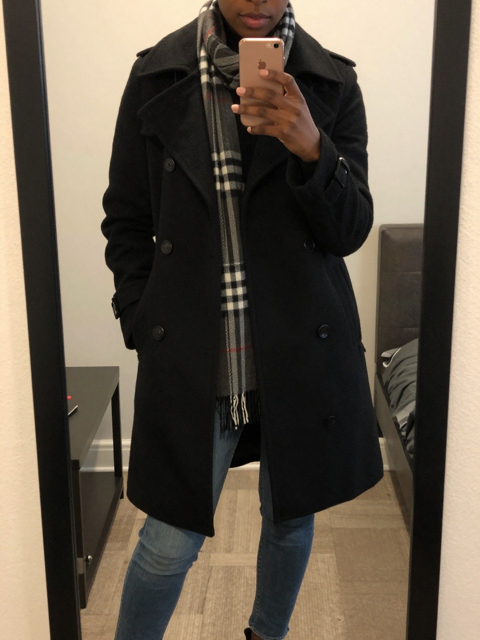 iPhone mirror selfie of a Black woman wearing a black peacoat with a plaid scarf and blue jeans, standing casually with her phone at her side, cropped at NECK so face doesn't show, in a city apartment, warm moody lighting, iPhone photo quality.