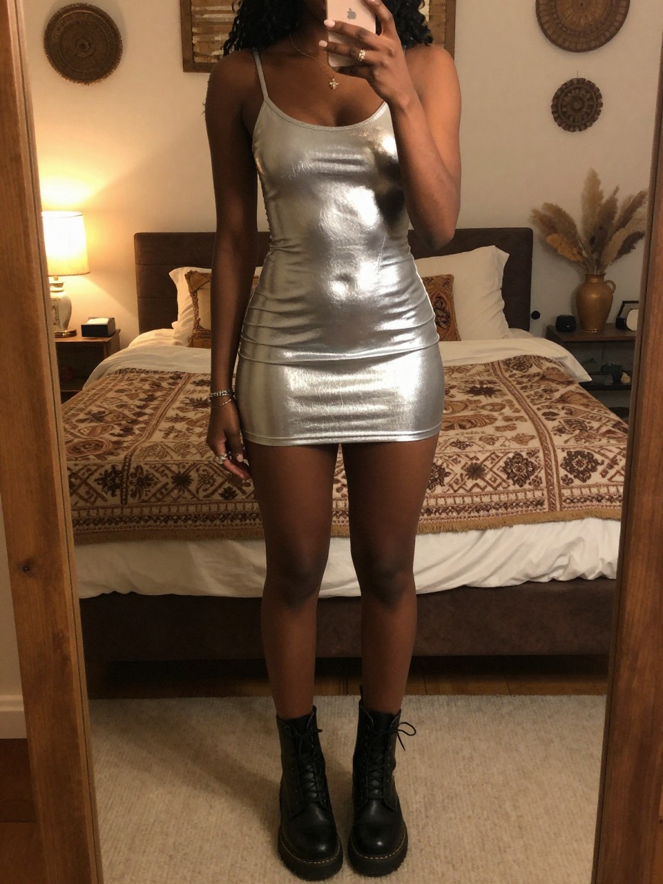 iPhone mirror selfie of a black woman in a silver metallic dress and combat boots, one hand behind head, cropped at NECK so face doesn't show, in a bohemian-themed bedroom with warm moody lighting, iPhone photo quality.