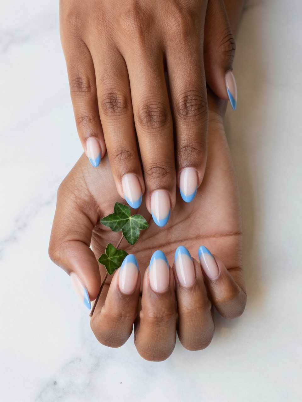 Close-up, top-down view of black feminine hands with long, natural almond nails on a marble surface. The nails have a powder blue French tip. A single strand of ivy with three small leaves trails from the tip down the nail. The look is clean, green, and graceful.