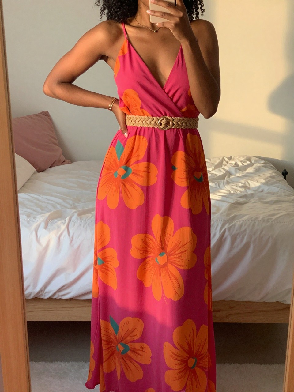 iPhone mirror selfie of a black woman wearing a fuchsia and orange floral maxi dress with a woven belt, posing with one hand on her hip, cropped at NECK so face doesn't show, in a feminine-themed bedroom, evening golden hour light, iPhone photo quality.