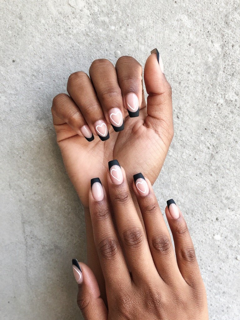 Top-down view of black woman hands with a matte black French manicure, posed against a concrete wall. A simple white heart outline is centered on each nude nail bed. The composition is sharp, graphic, and urban-chic.