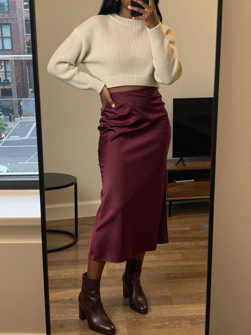 iPhone mirror selfie of a black woman wearing a cream cropped ribbed sweater with a high-waisted burgundy satin midi skirt and brown block-heel ankle boots, one hand on her hip, cropped at NECK so face doesn't show, in a city apartment, warm moody lighting, iPhone photo quality.