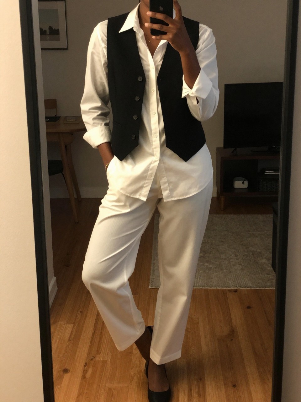 iPhone mirror selfie of a black woman wearing a black vest over a white button-down with trousers, standing with her weight on one leg and hands in pockets, cropped at NECK so face doesn't show, in a city apartment, warm moody lighting, iPhone photo quality.