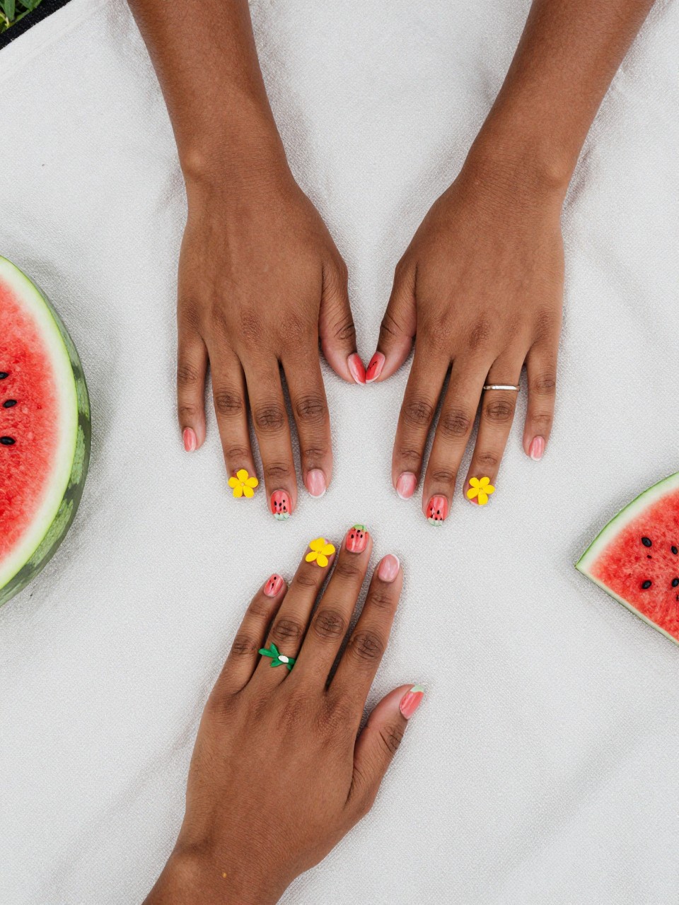 Top-down view of black woman hands on a picnic cloth. The nails are designed as watermelon slices. The nails also feature a simple yellow watermelon blossom on a green vine. The setting is a picnic blanket in the park.