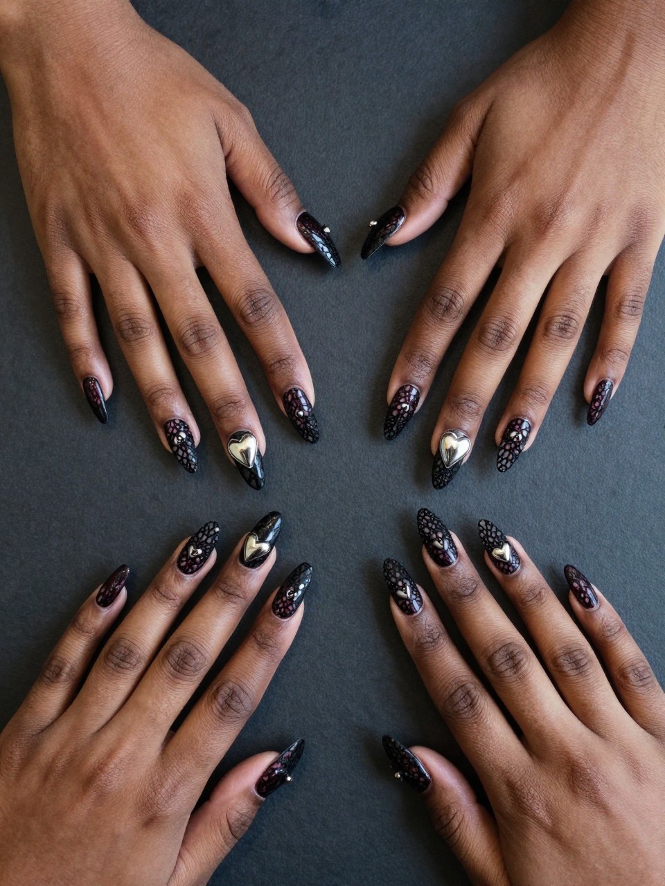 Bird's-eye view of black feminine hands laid on a table. The nail designs feature a detailed black lace pattern over oxblood. A single chrome heart sits in the center of the lace motif on each nail. The mood is gothic romance.